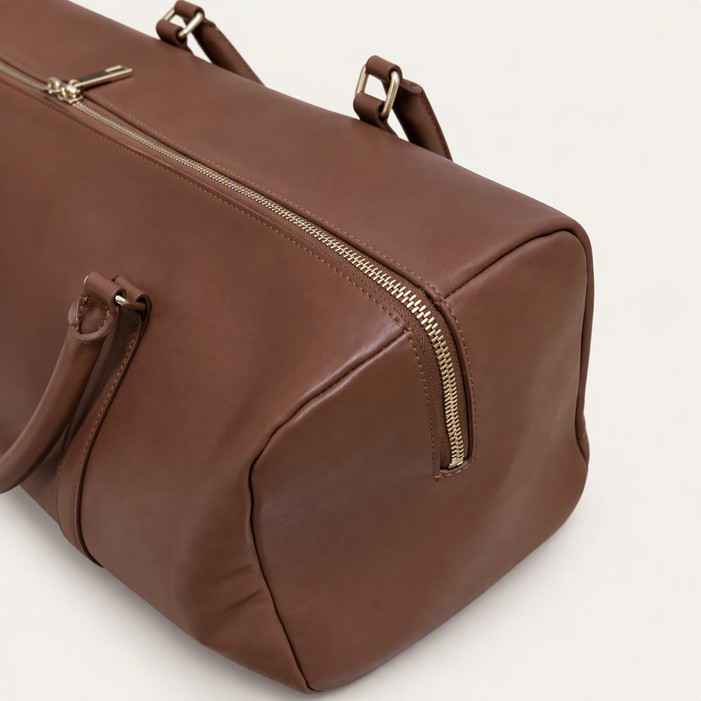 A brown leather duffel bag with gold-tone zippers and handles, shown at an angle on a light off-white background.