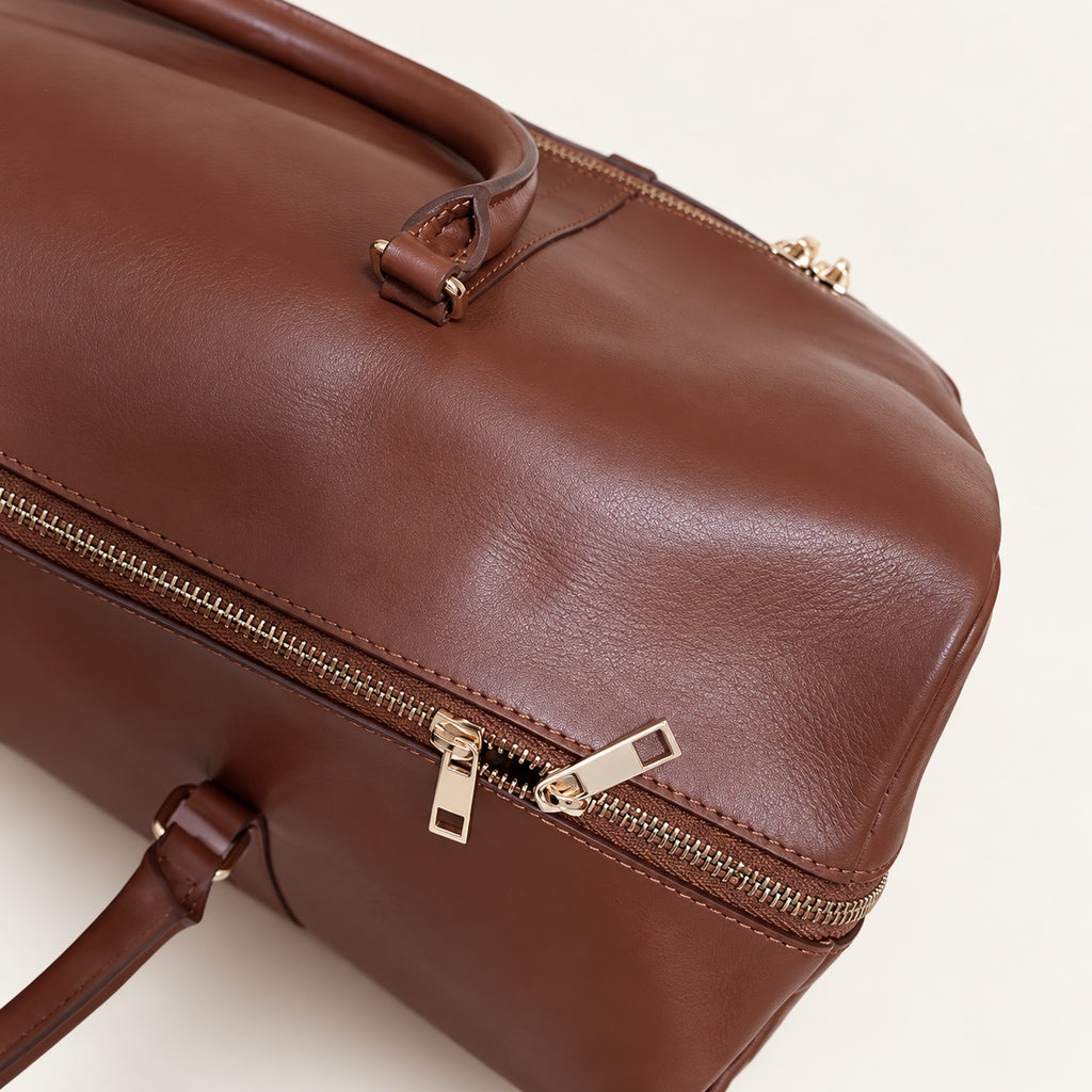 A close-up of a brown leather duffel bag with gold-tone zippers and a matching leather handle, shown against a plain off-white background.