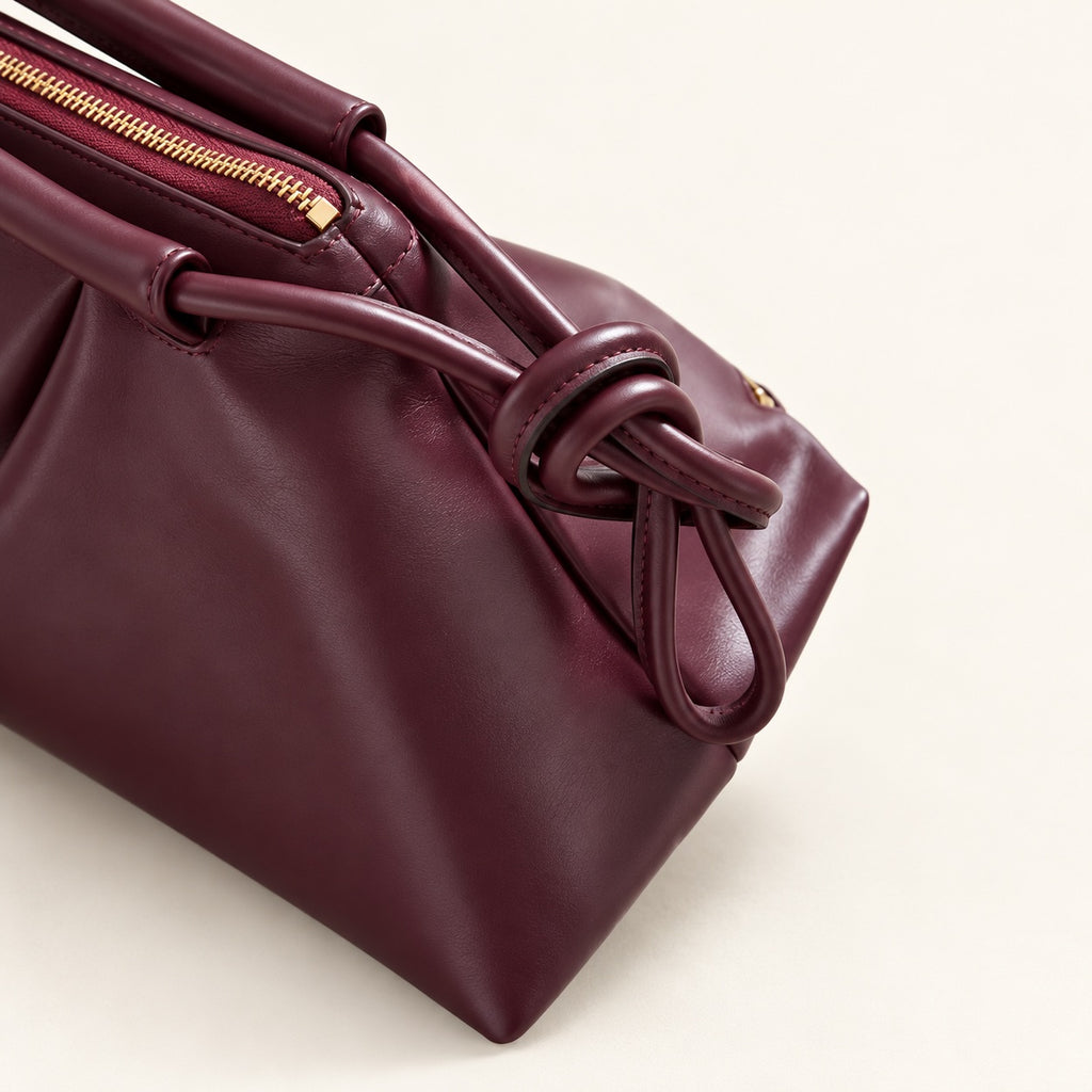 A close-up of a structured leather handbag in deep burgundy, featuring a gold zipper and a knotted leather handle detail, set against a clean off-white background.