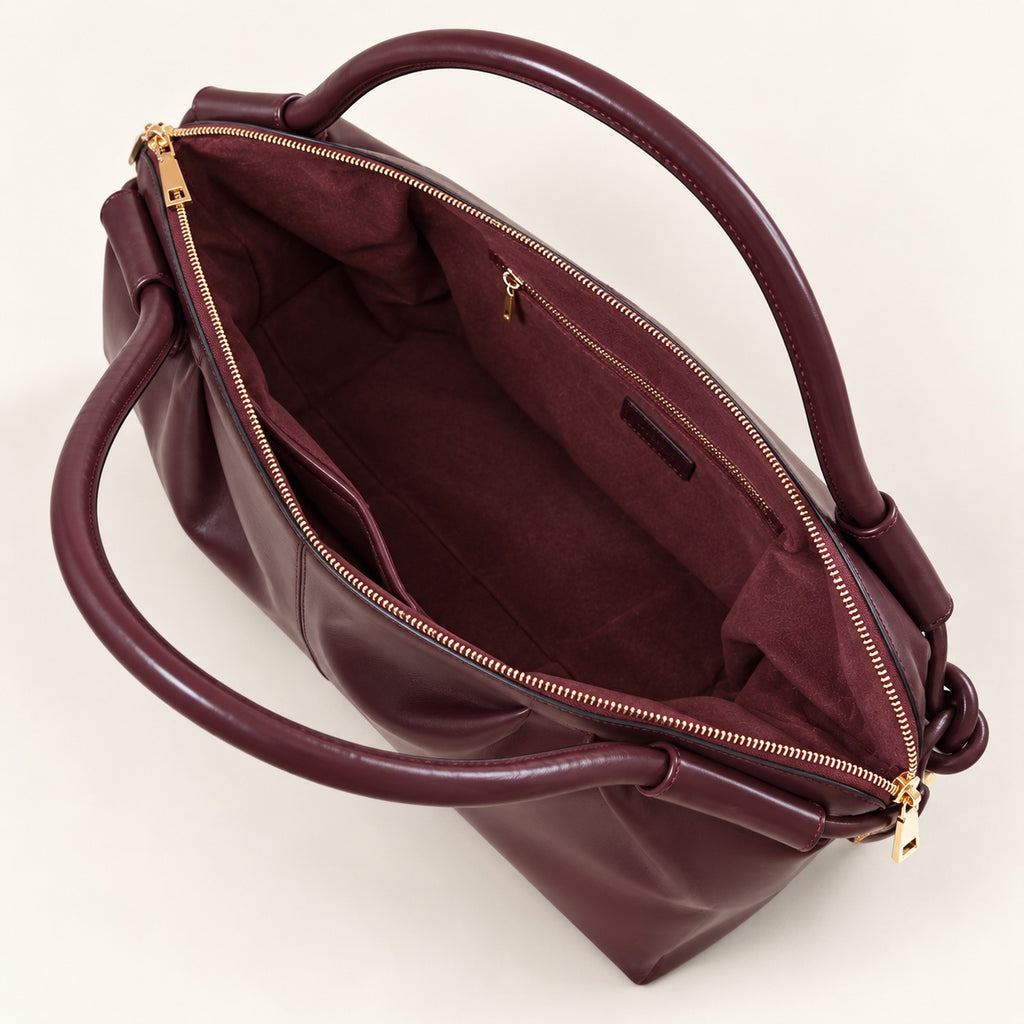 A top-down view of a dark burgundy leather handbag with gold-tone hardware, showing its interior lining, zippered compartments, and double top handles against a light off-white background.