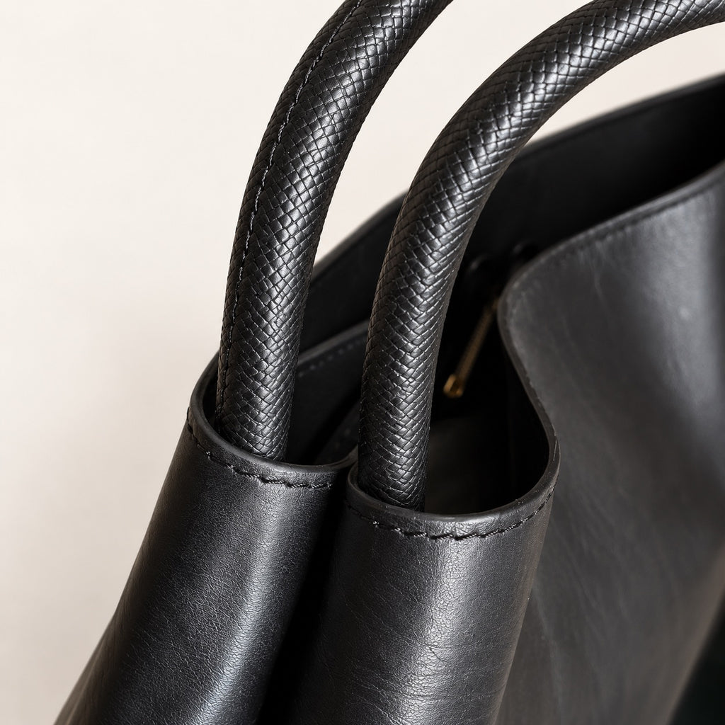 Close-up detail of a black leather tote bag with braided double handles and visible stitching, set against a plain light background.