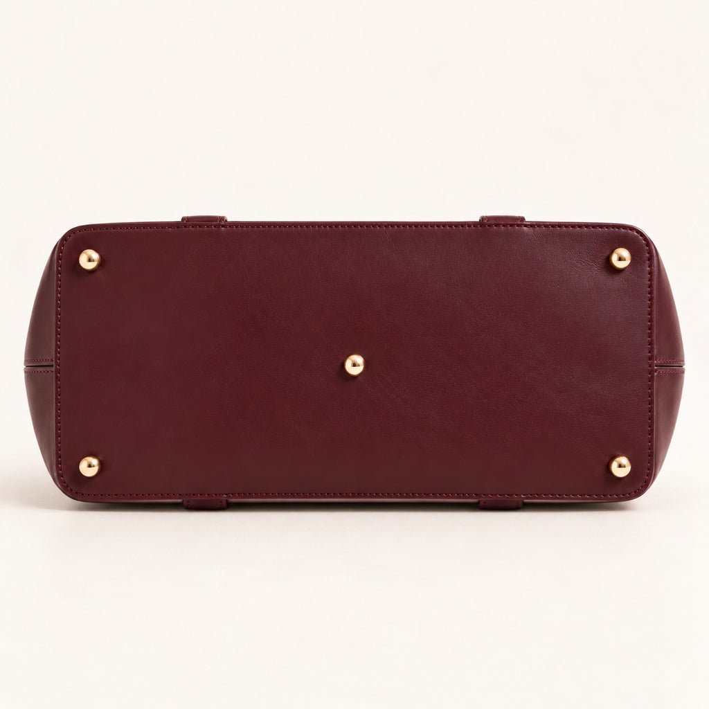 The bottom view of a burgundy leather clutch or handbag, showing a smooth leather surface with four gold-tone metal studs on the corners and a central gold stud, set against a plain off-white background.